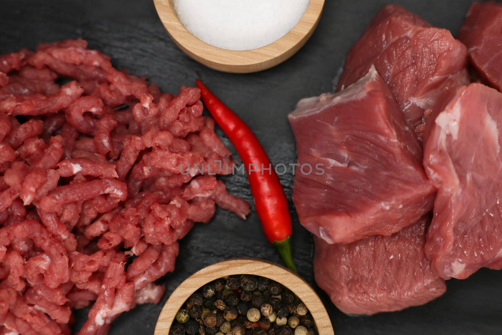 A close-up view from above of chunks of beef meat and red minced meat with streaks of fat lying on a black background with spices and salt in wooden bowls and red hot peppers in the center. The concept of cooking, shredding meat, diet, natural products. High quality photo
