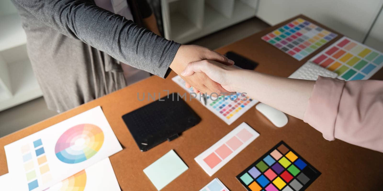 A close-up shot of graphic designers shaking hands, surrounded by color palettes and design materials on a collaborative workspace desk.
