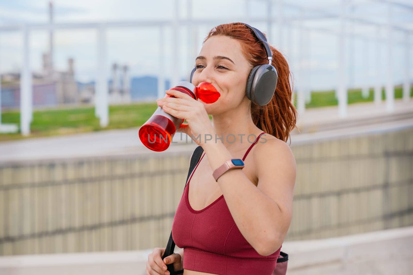 Athletic red-haired woman wearing sportswear and headphones, drinking water after exercise. She carries a gym bag and stands in a modern outdoor urban environment under a cloudy sky.