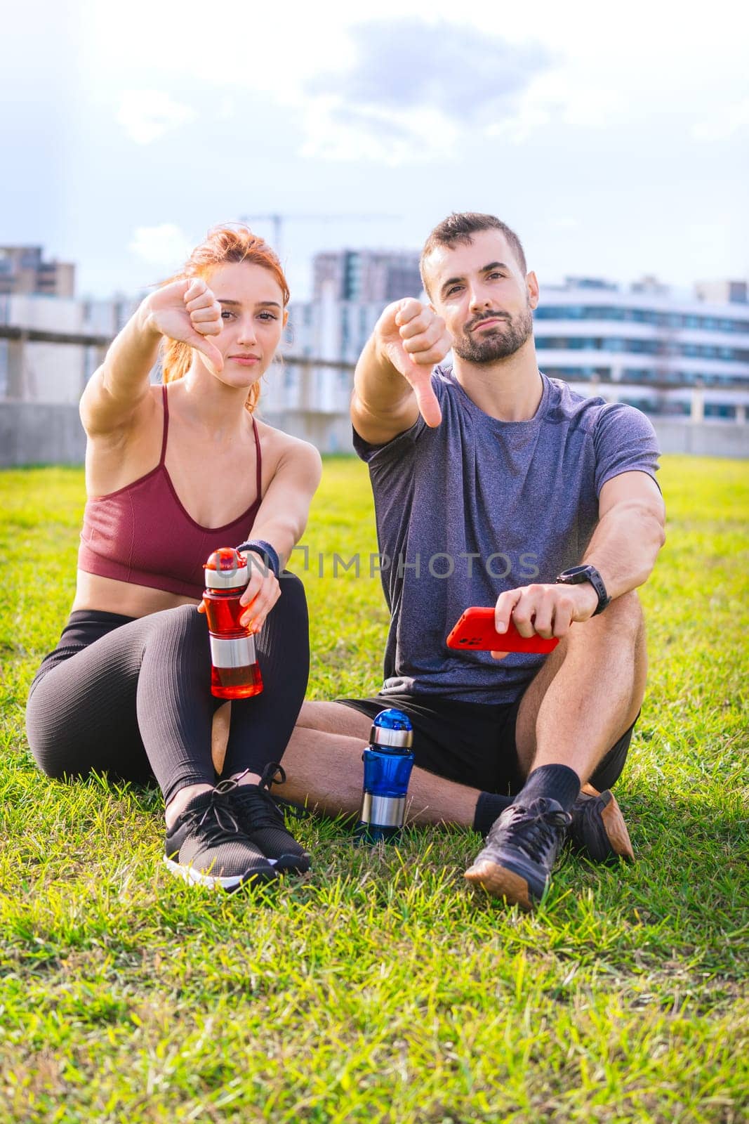 A fit man and woman sitting on the grass after a workout, holding water bottles and pointing at the camera. They appear relaxed and playful, enjoying a sunny outdoor setting in the city.