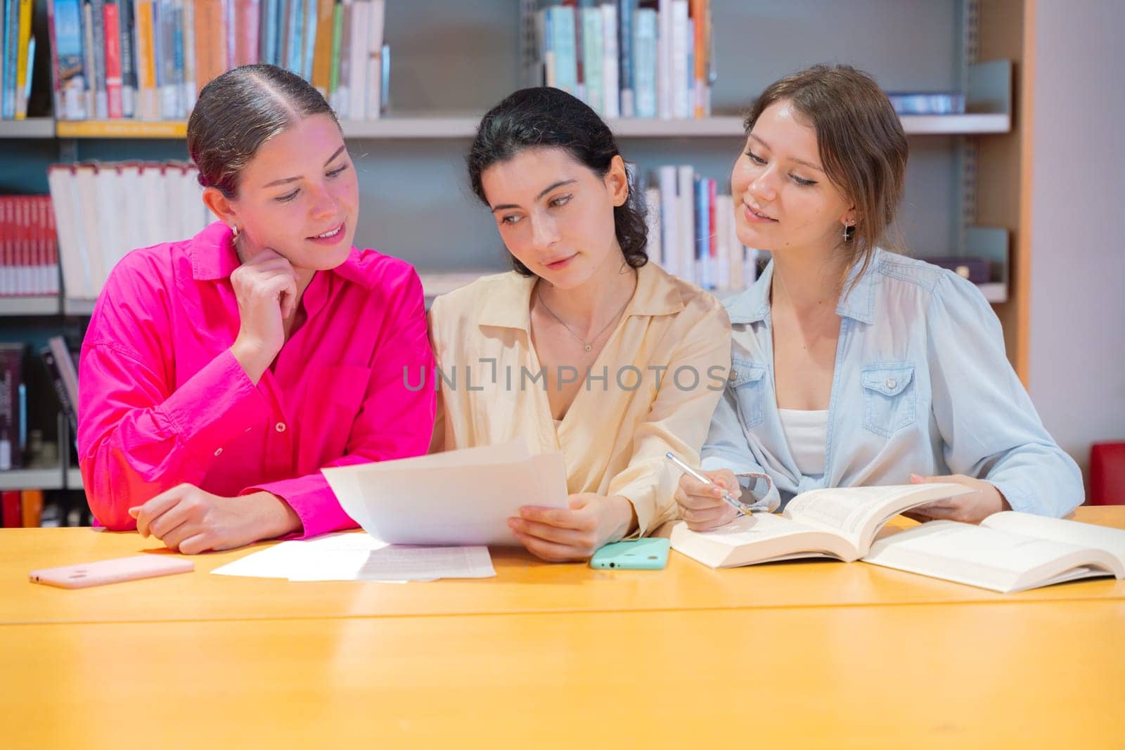 Three young women collaborating at a library table, studying and discussing documents. They are engaged in learning, taking notes, and exchanging ideas in a focused academic environment.