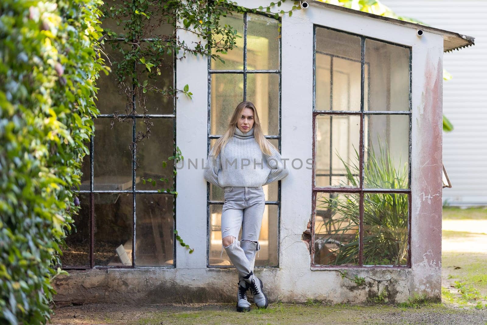 Young woman is standing in front of an old greenhouse, leaning on the wall with her arms behind her back