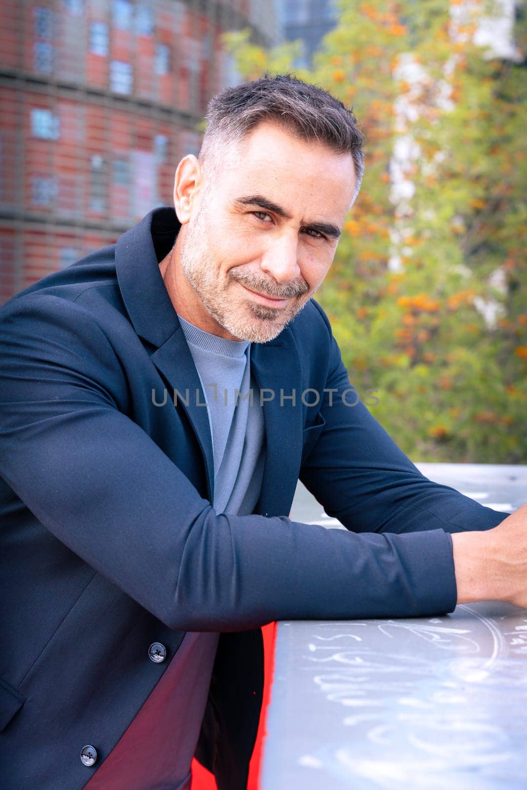 A middle-aged businessman in a stylish suit leaning on a railing and smiling. The modern city background enhances his confident, approachable, and professional demeanor.
