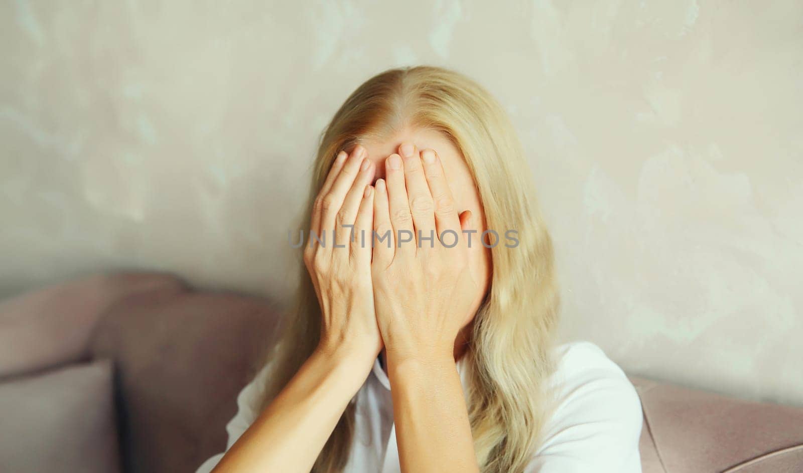 Upset sad crying woman covering her eyes and face with her hands while experiencing mental or physical suffering in room at home