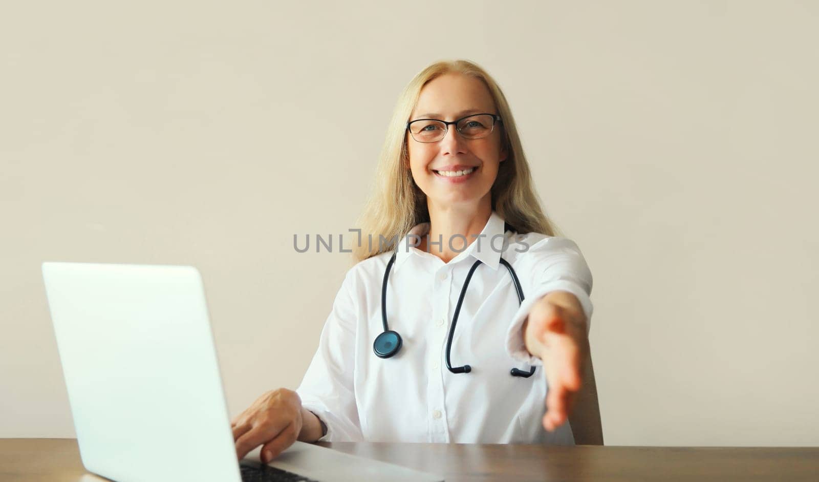 Happy middle-aged woman doctor working with laptop computer while sitting at desk in medical clinic office