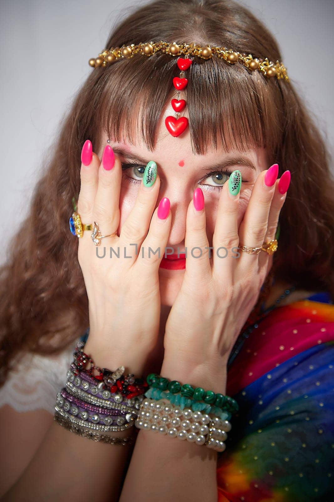 A blonde woman showcases a colorful sari while playfully covering her face with her hands, adorned with jewelry, during a lifestyle photoshoot.