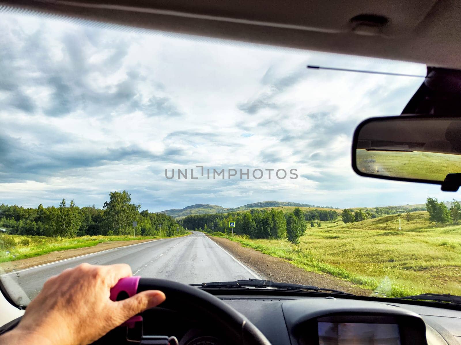 view through car windshield of nature landscape with green field, mountains, white clouds. Hand of woman on steering wheel. Female traveler