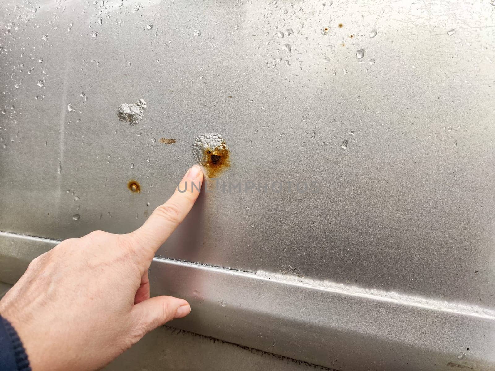 A person examines rust and scratches on silver car parked in a bright outdoor setting, focusing on the damaged area