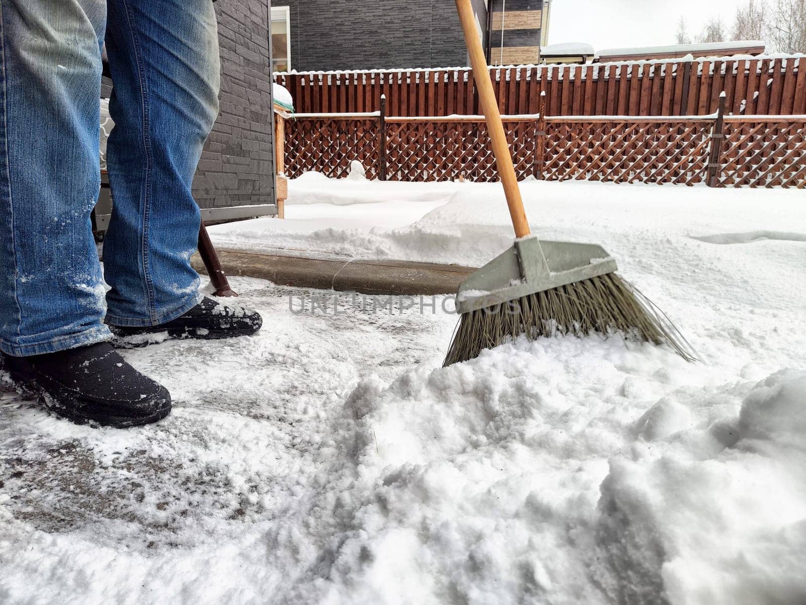 Individual uses a broom to clear snow from the driveway in a residential neighborhood during winter, showcasing heavy snowfall accumulation.