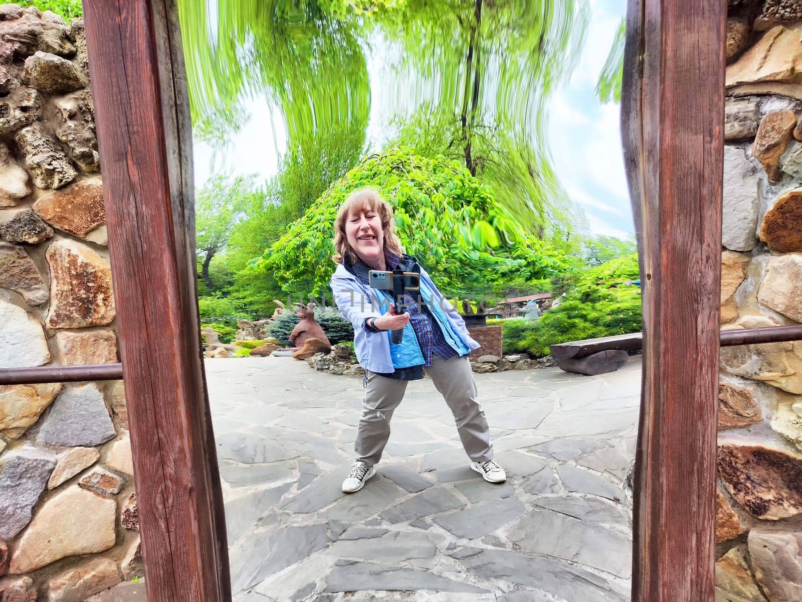 A modern blonde woman playfully takes a selfie in a curved mirror at a peaceful park during her adventure.
