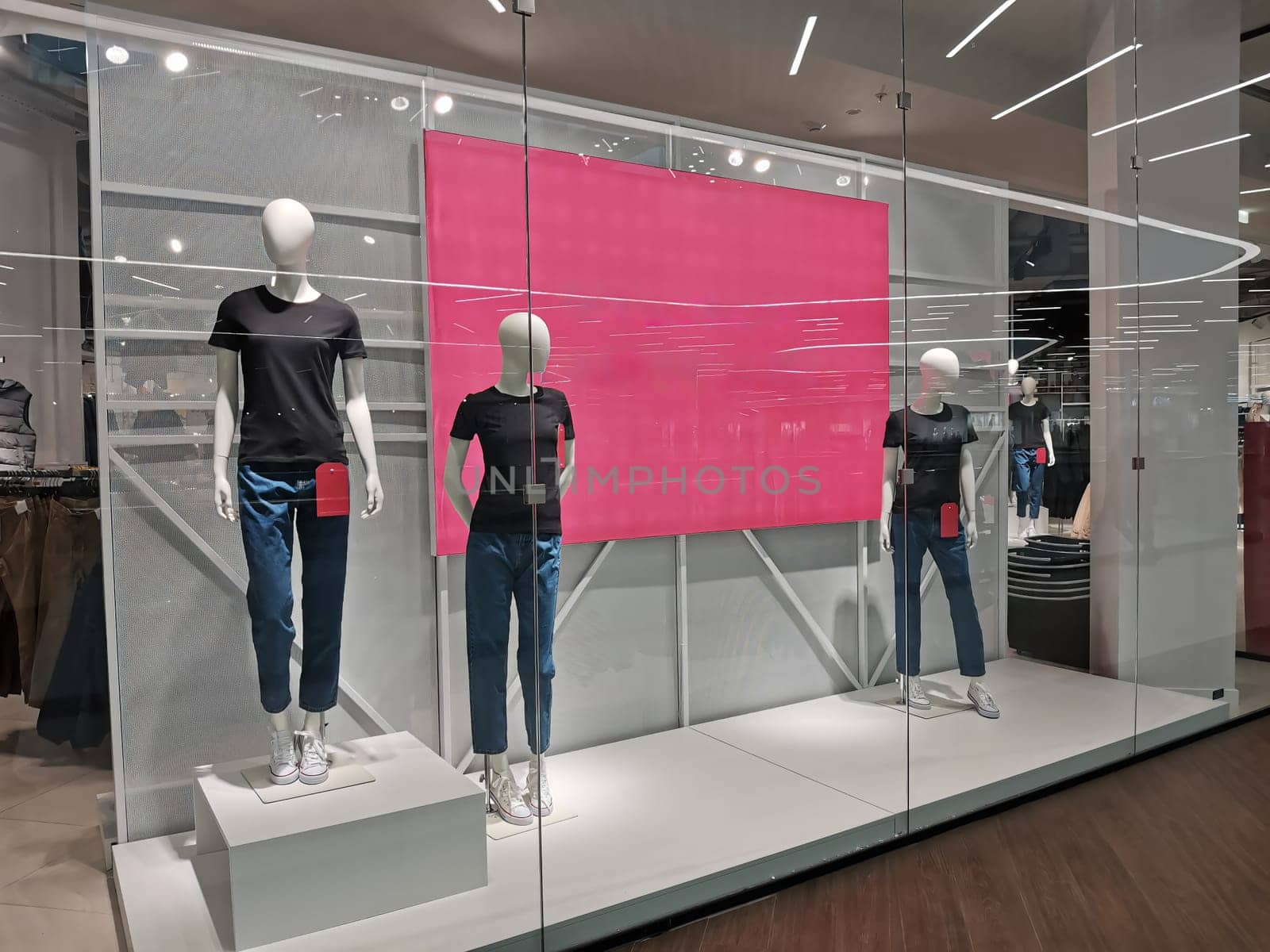 Window bright display with three mannequins wearing jeans and black t-shirts with empty red pink banner. Sale concept. Boutique retail window with copy space