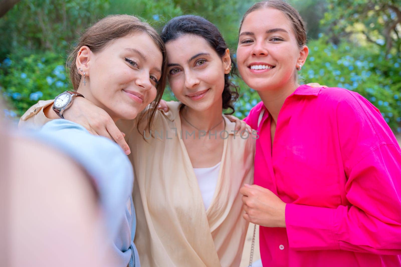 Three smiling women posing for a selfie in a lush green park. They embrace each other, expressing happiness and friendship. The vibrant colors and natural background create a joyful atmosphere.