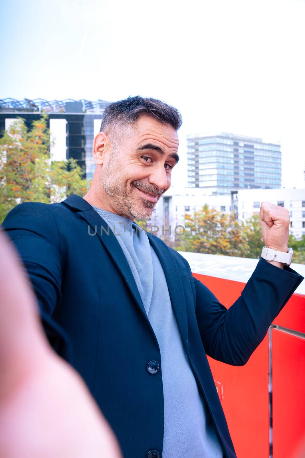 Happy middle-aged man in a blazer taking a selfie with a cheerful expression. He raises his fist in excitement. The background features a modern urban setting with buildings and trees.