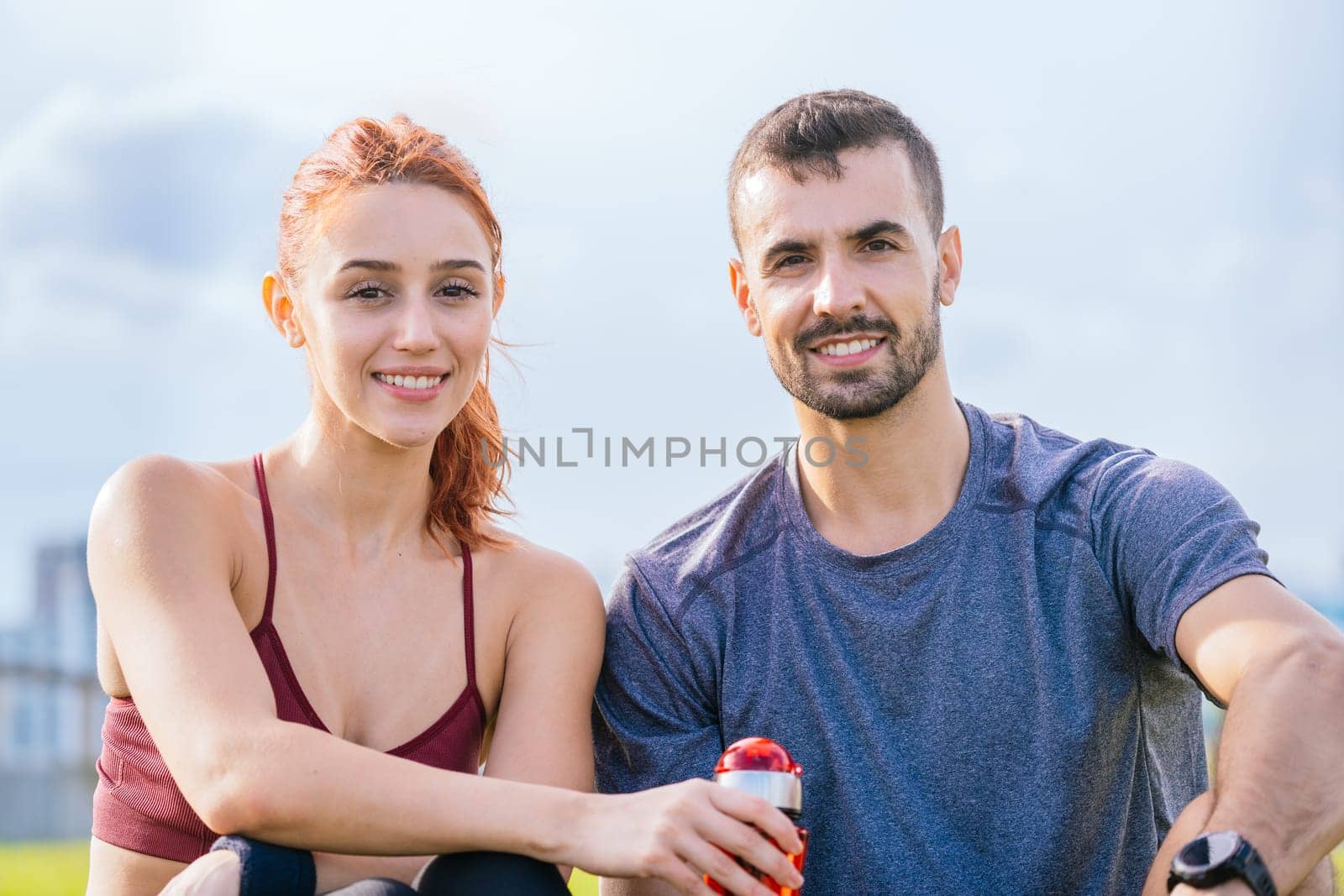 Young man and woman in sportswear sitting outdoors, smiling and relaxing after exercise. They are holding a drink, enjoying the fresh air in a park with a bright sky in the background.