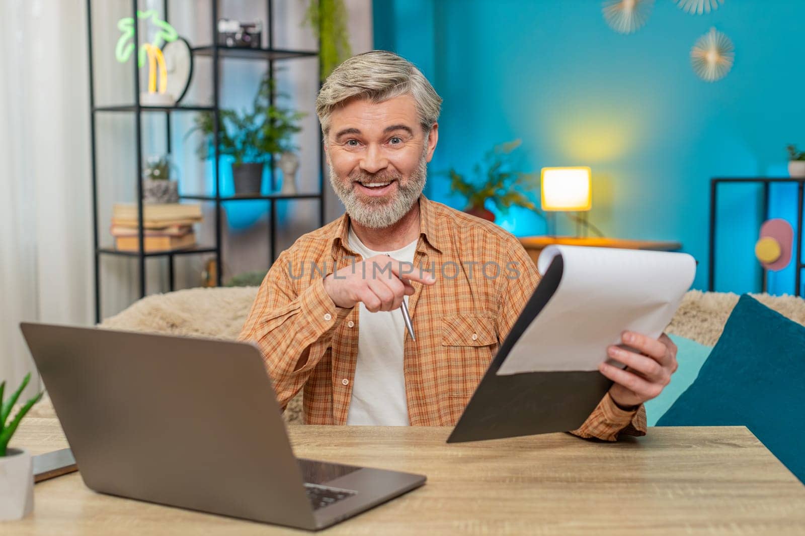 Caucasian mature man sitting at home table reviewing important documents and using laptop. Middle-aged guy smiles with satisfaction from successful work, demonstrating productivity and accomplishment