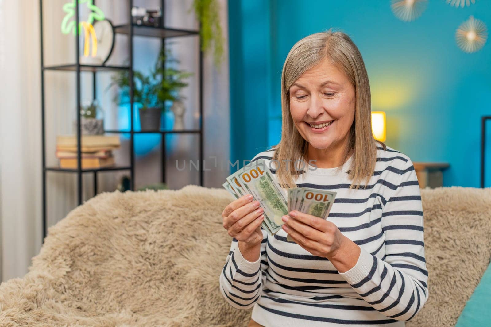 Happy Caucasian mature woman counts money dollar cash in hands after winning lottery bonus or receiving a gift. Grandmother smiles broadly sitting on home sofa, celebrating financial victory salary