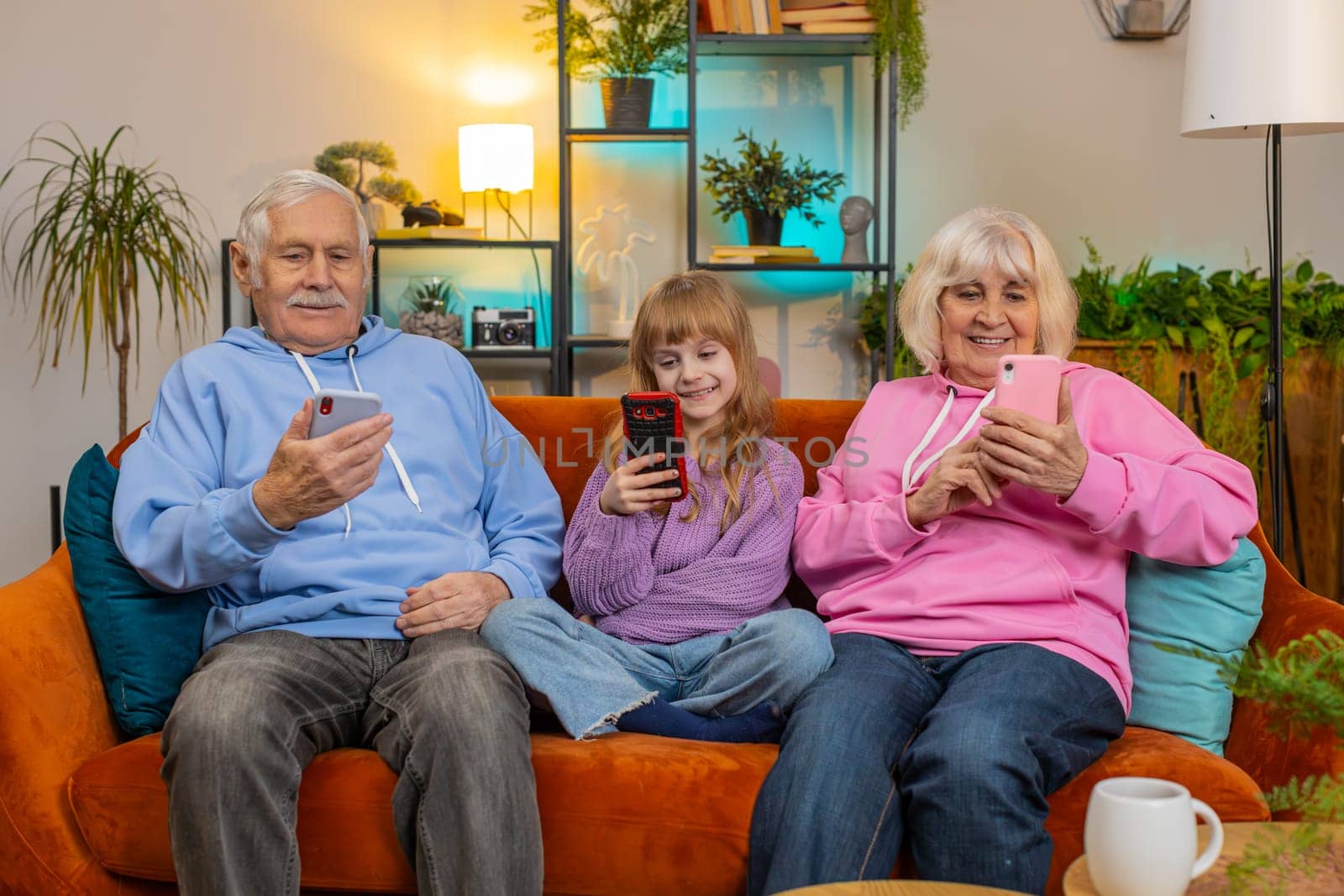 Caucasian grandfather, grandmother and granddaughter using smartphones in living room at home. Smiling girl and senior couple spending leisure time sitting on sofa. Playing online games, social media.