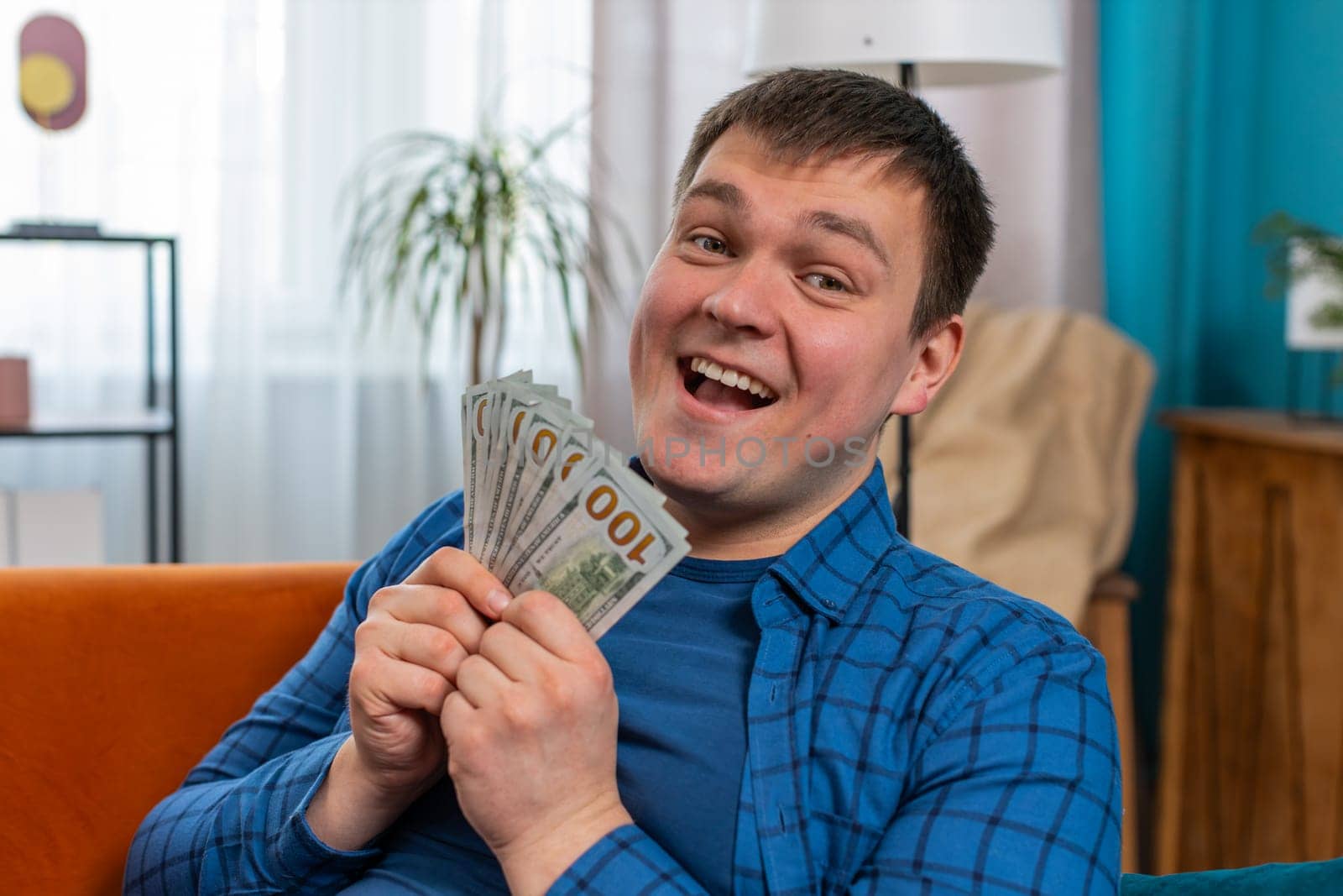 Happy rich Caucasian young man cheering up with high money profits, lottery game win, satisfied of wealth income earnings salary sit on home room couch sofa full of dollar cash banknotes in apartment.