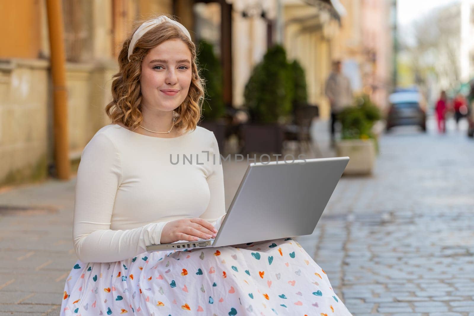 Woman freelancer sitting on city street using laptop working online distant job browsing website chatting outdoors during break. Girl looking at camera white sends messages watching movies at notebook