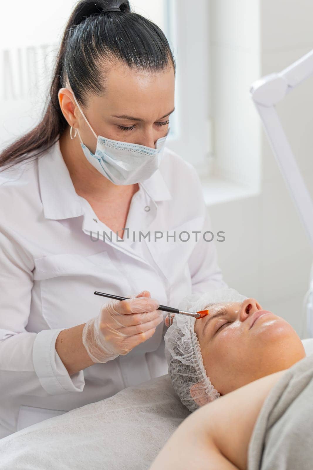 Face peeling mask, spa beauty treatment, skincare. Beautician applying vitamins moisturizing cream on young woman face for rehydrate face skin, anti aging cosmetic procedure in beauty salon. Vertical.