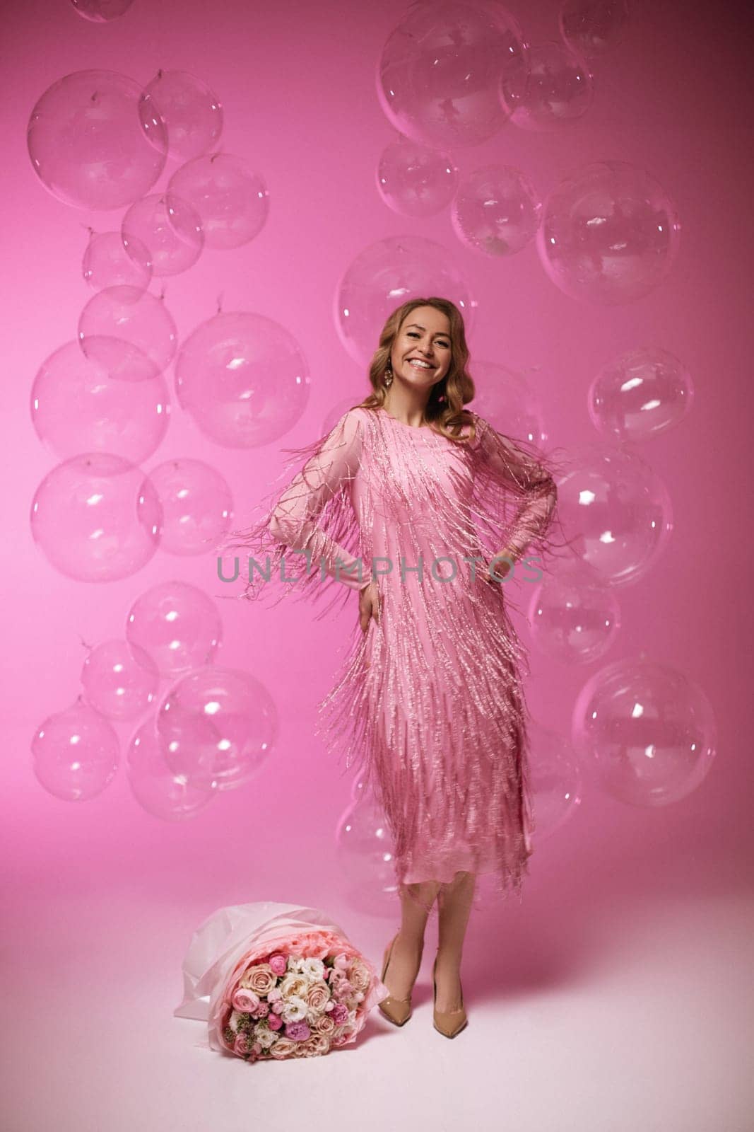 Beautiful woman in amazing long ball dress is holding pink balloons and is posing on pink background.