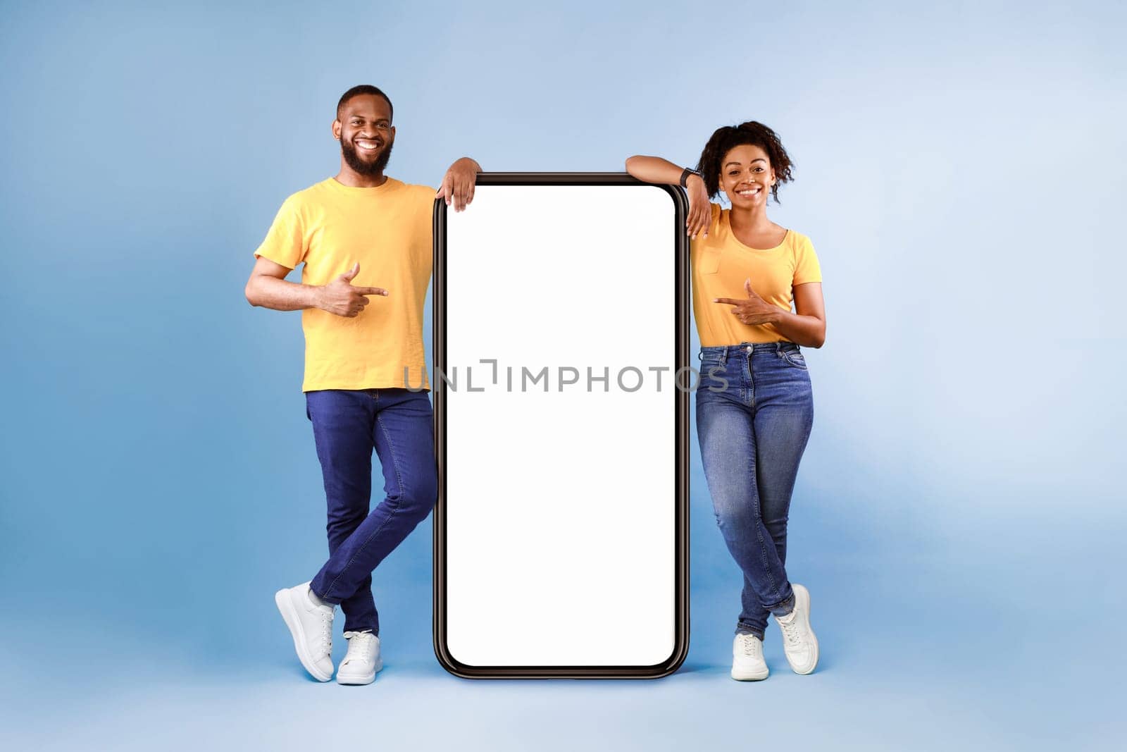 Great mobile app. African American couple pointing at giant smartphone with mockup, promoting application or website, advertising product or service, blue background. Full length