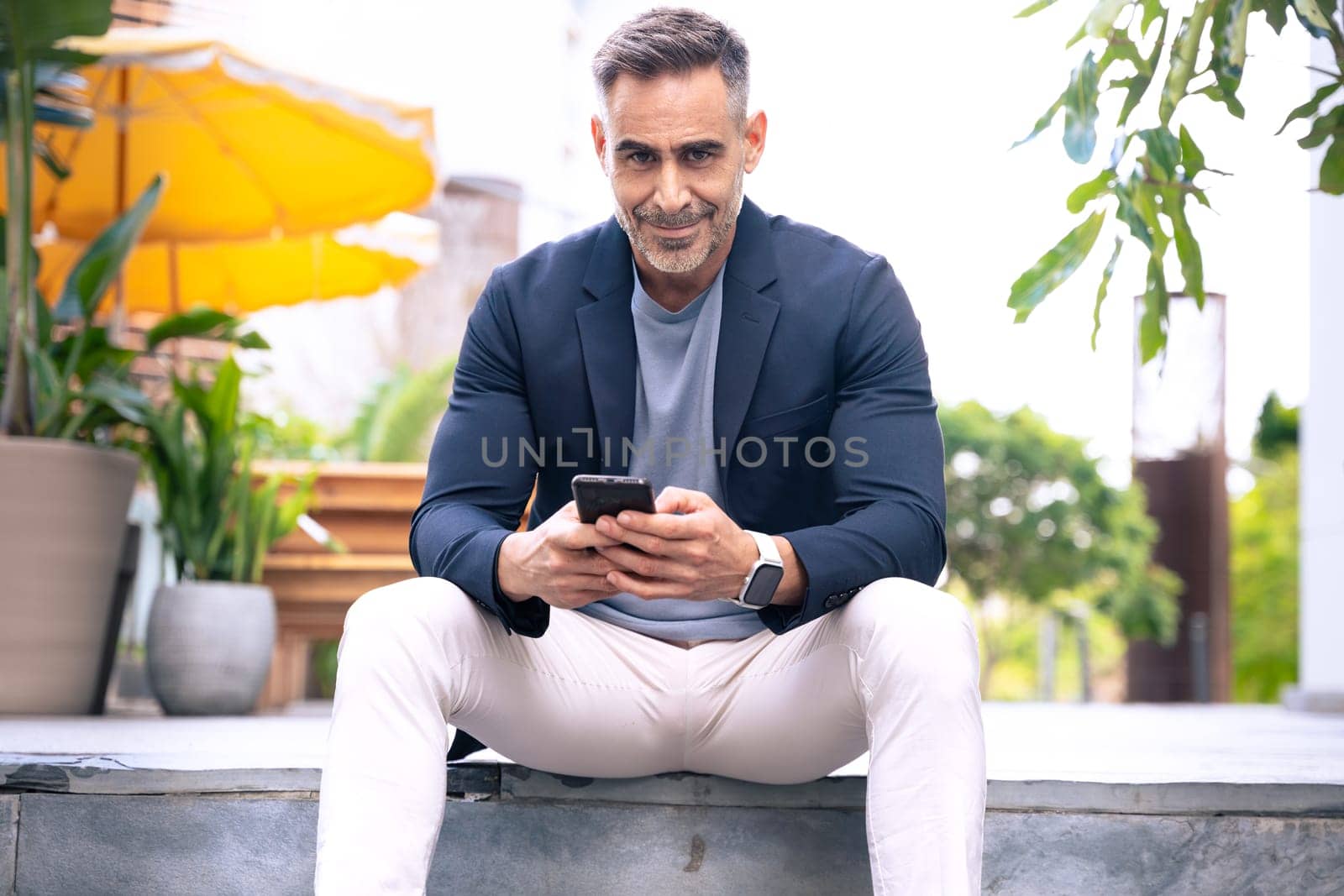 Happy middle-aged man in a blazer sitting on steps while using his smartphone. He has a relaxed and focused expression. The background features an outdoor urban environment with greenery and seating.