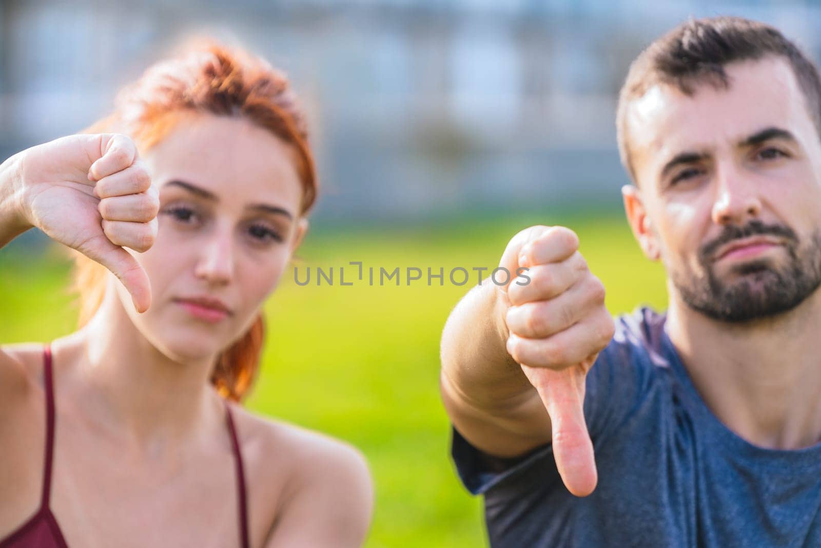 Young man and woman in sportswear sitting outdoors on the grass, looking displeased and showing thumbs down. They are holding water bottles, resting after exercise in an urban park setting.