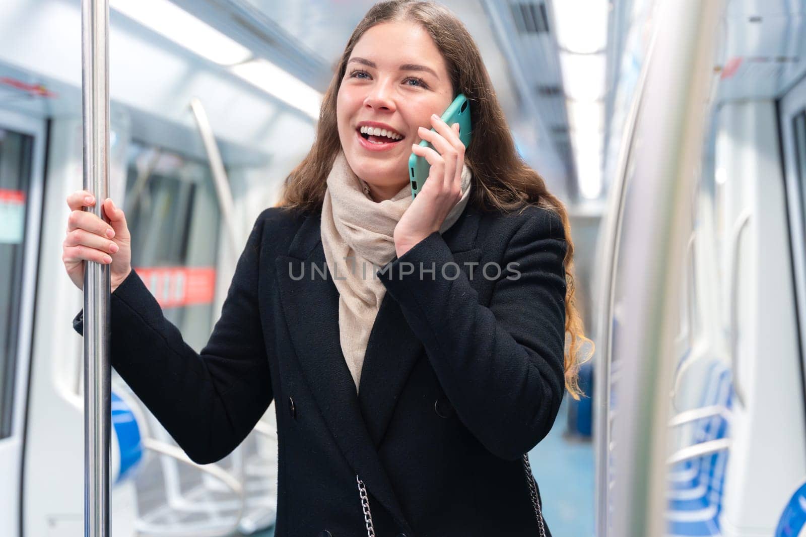Cheerful young woman in a black coat and beige scarf talking on her smartphone while standing inside a modern subway train. She smiles brightly, holding onto a metal pole during her commute.