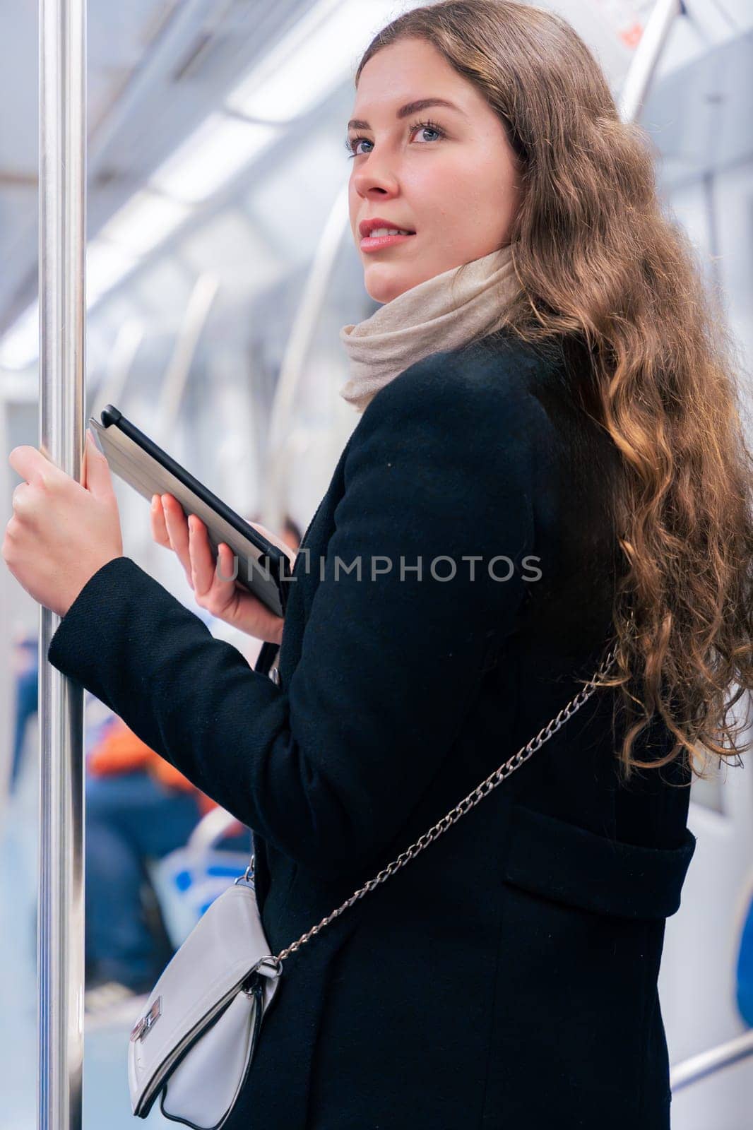 Stylish young woman in a black coat and beige scarf reading on an e-reader while standing inside a modern subway train. She balances while holding onto a pole during her commute.