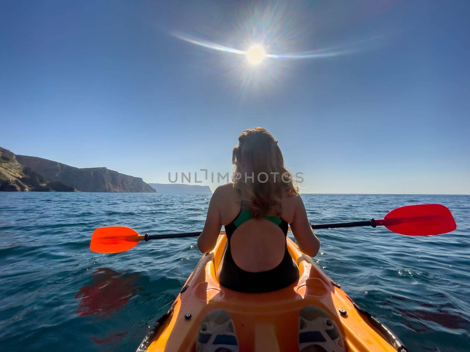 A woman in a swimsuit kayaks across a calm body of water, paddling with an orange oar. The sun shines brightly in the blue sky.