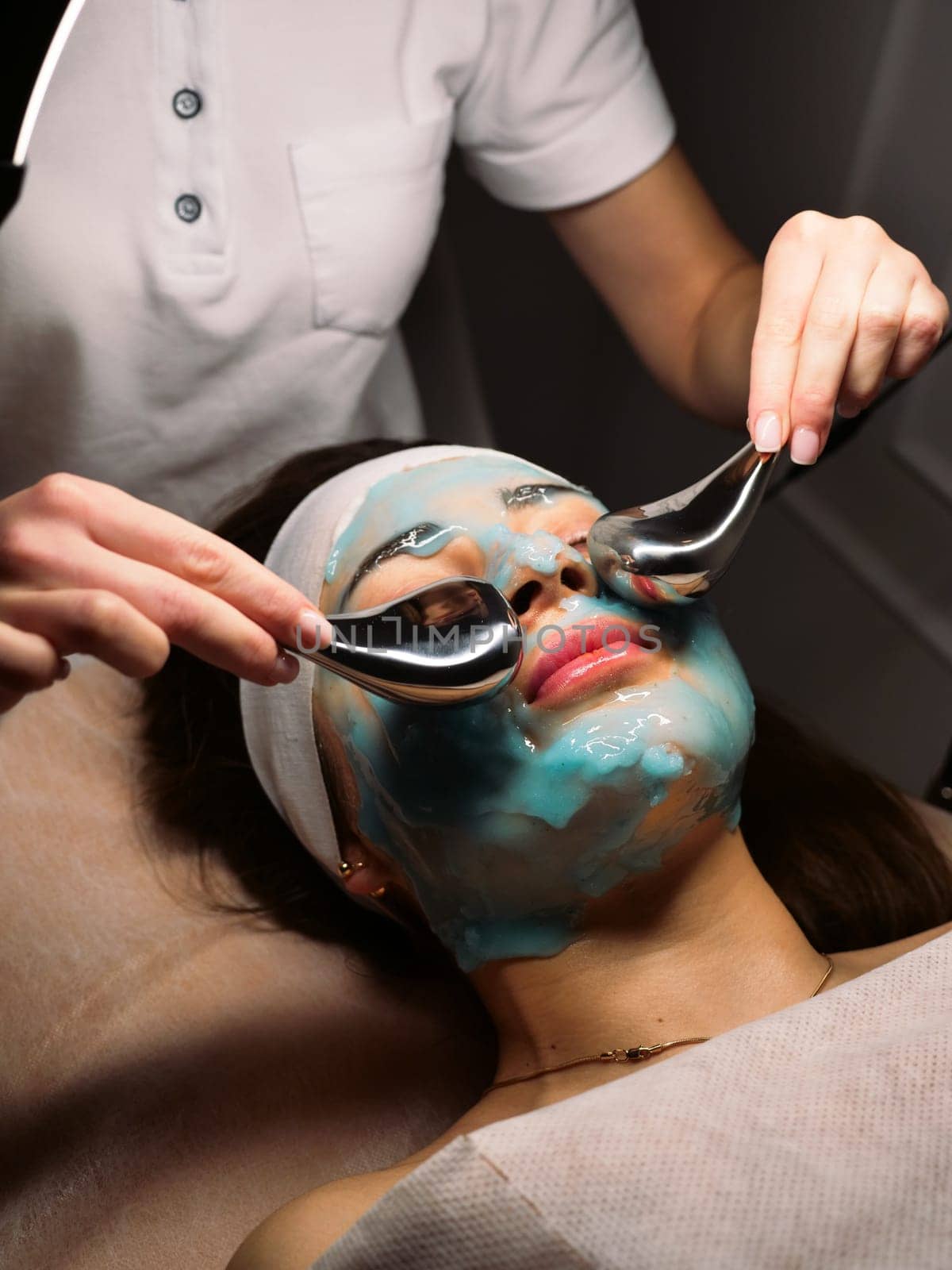 Cosmetologist does cryo massage with globules on a blue hydrogel alginate mask in the beauty salon, vertically, close-up. Woman Facial Skin & Body Care
