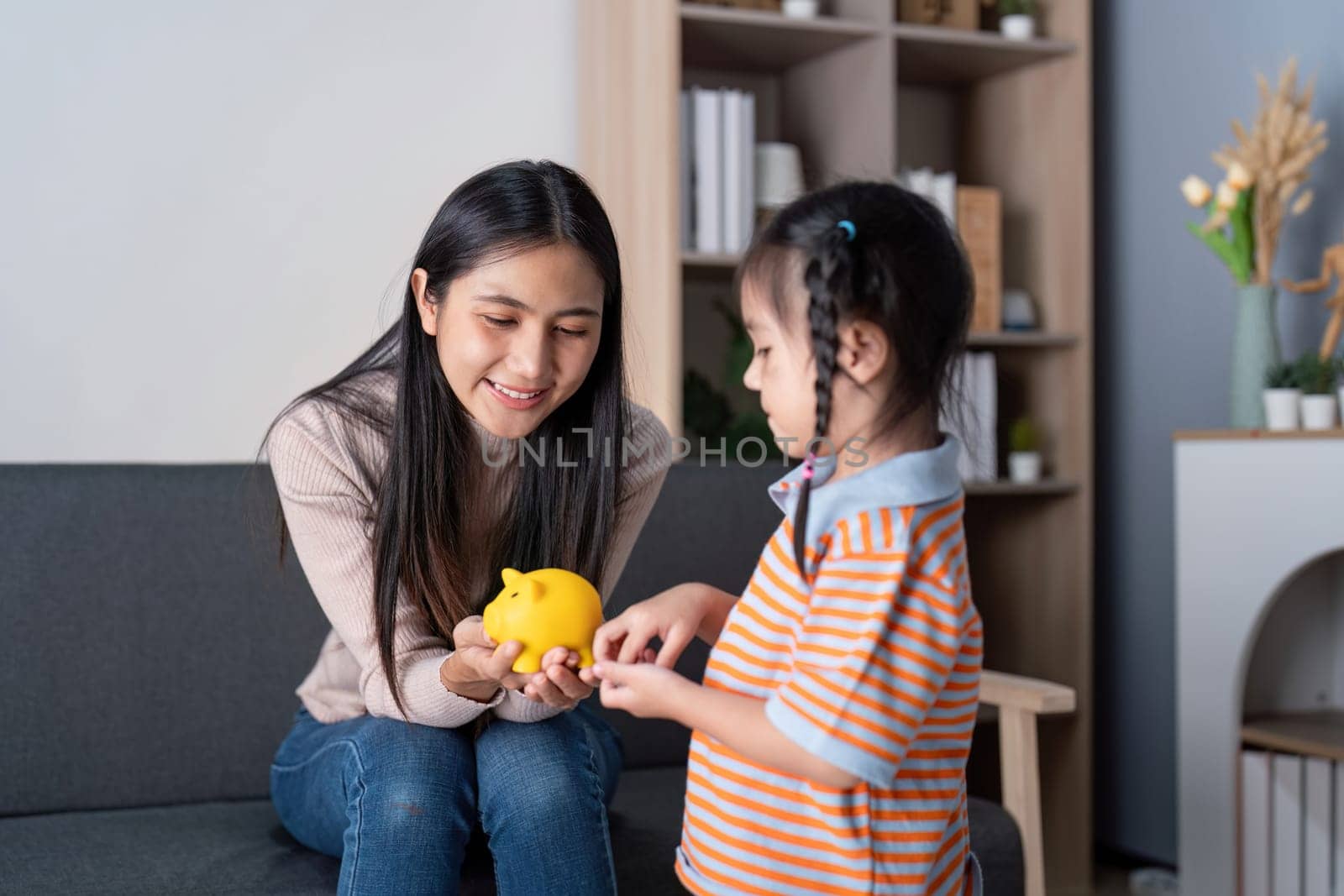 A mother joyfully teaches her daughter about saving money using a piggy bank in their inviting living room.