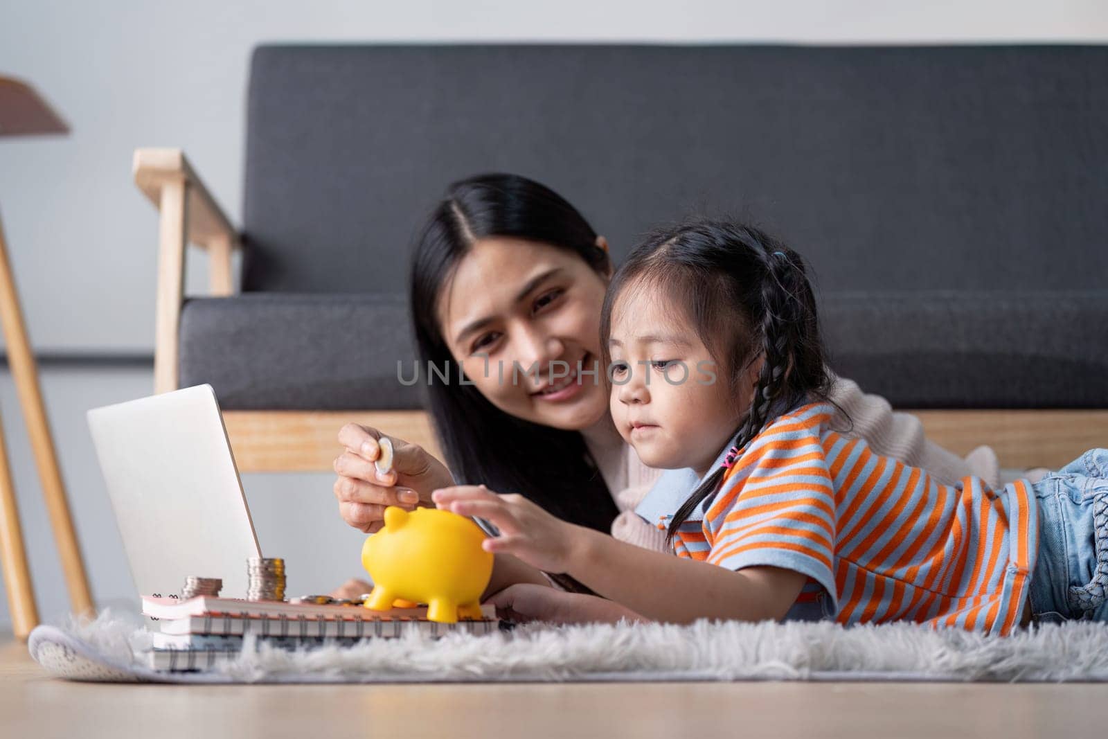 A diverse mother and her daughter enjoy a learning moment as they save money using a yellow piggy bank and a laptop, fostering financial literacy at home.
