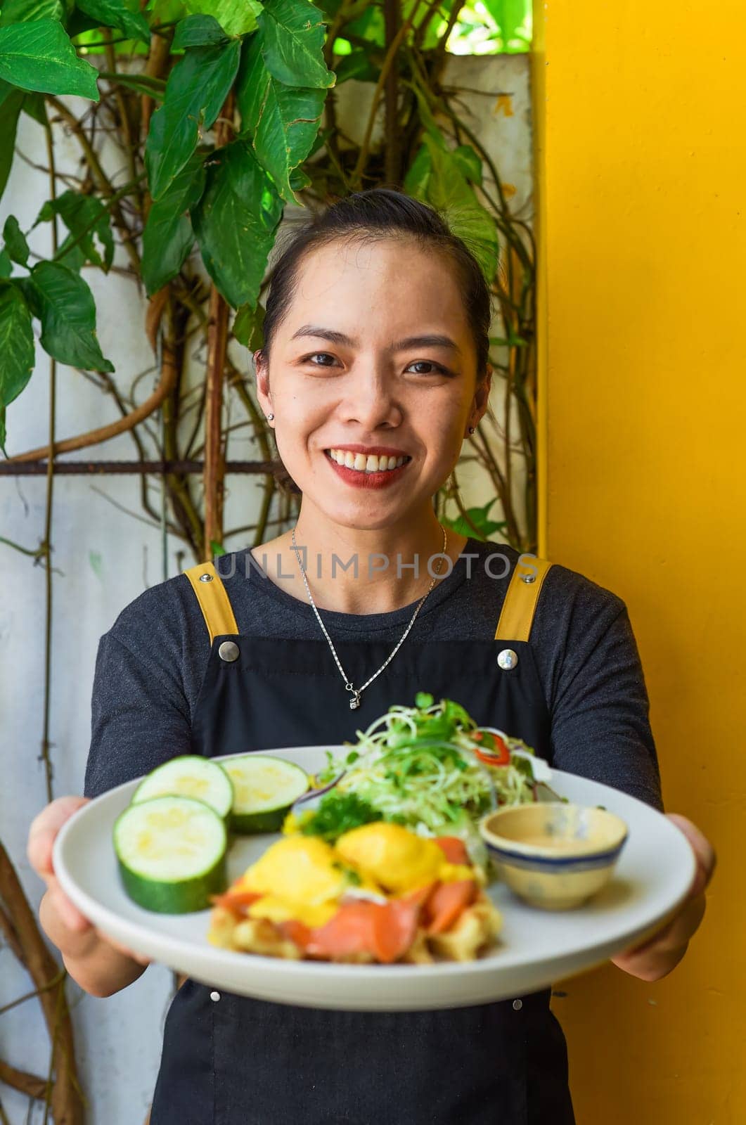Young vietnamese waitress holding a plate with eggs bennedict waffles with smoked salmon in restaurant