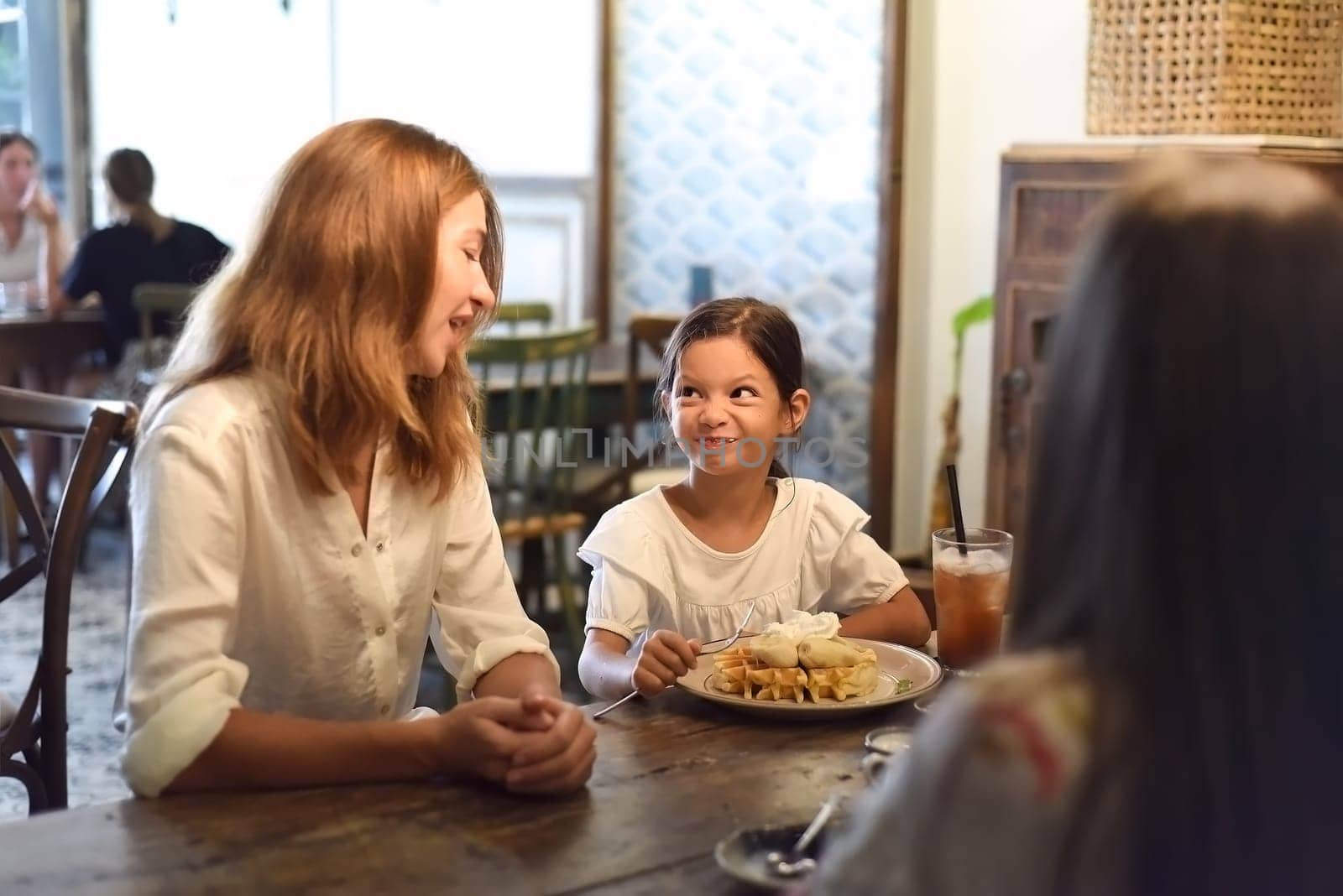 Little girl and her mother eating sweet belgian waffles with banana and cream in a cafe, daughter making a funny face