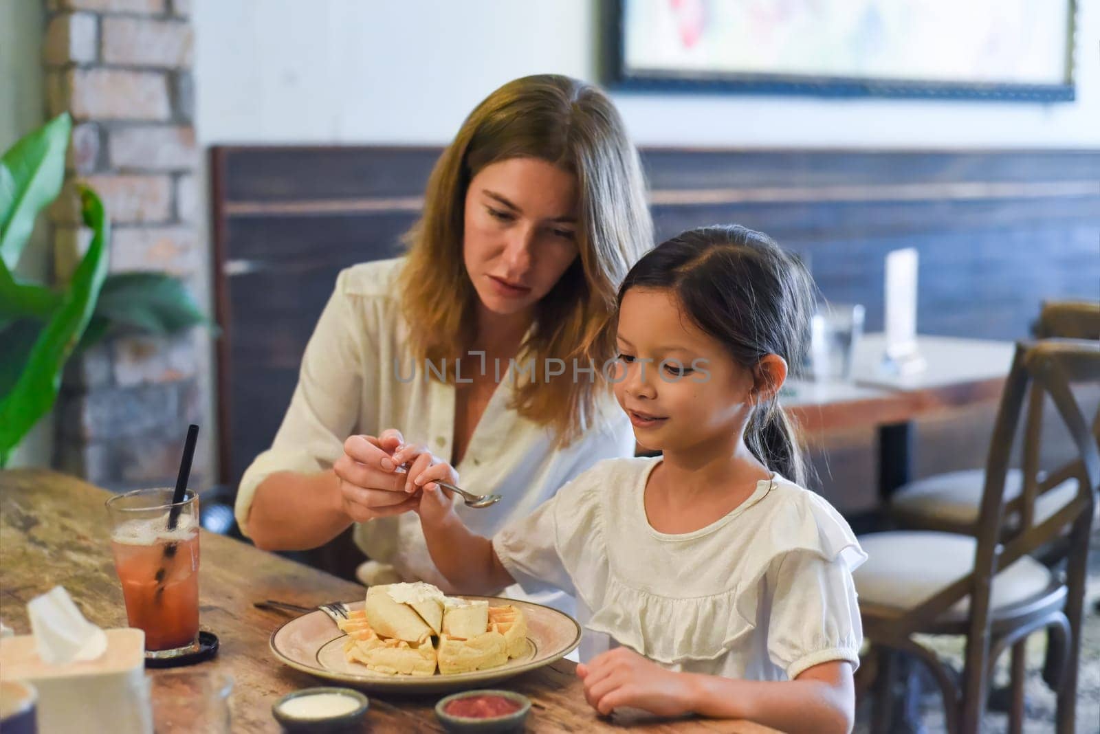 Little girl and her mother eating sweet belgian waffles with banana and cream in a cafe, daughter making a funny face