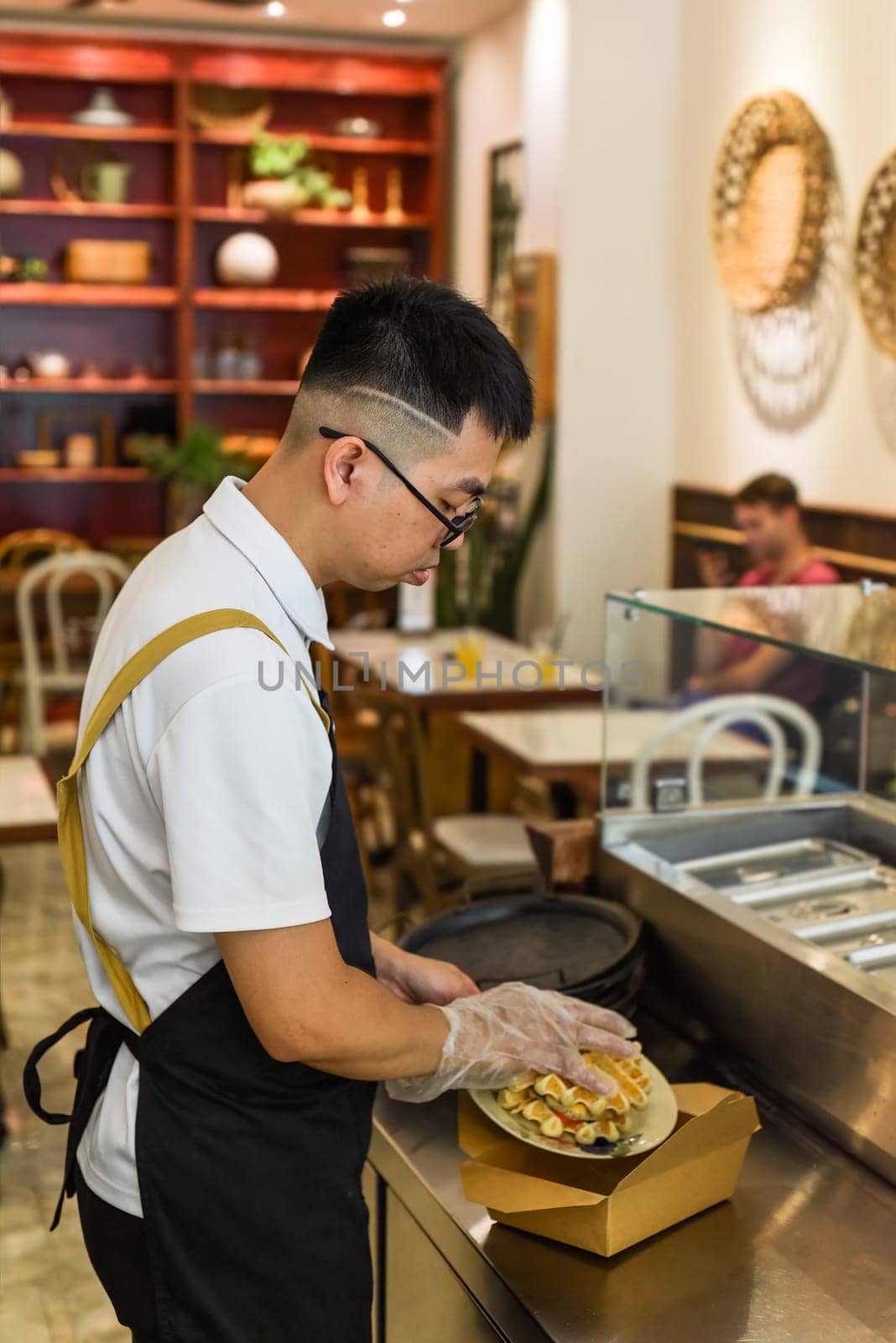 Vietnamese man chef in uniform holding fresh baked belgian waffles with chicken and putting them in paper box for take away