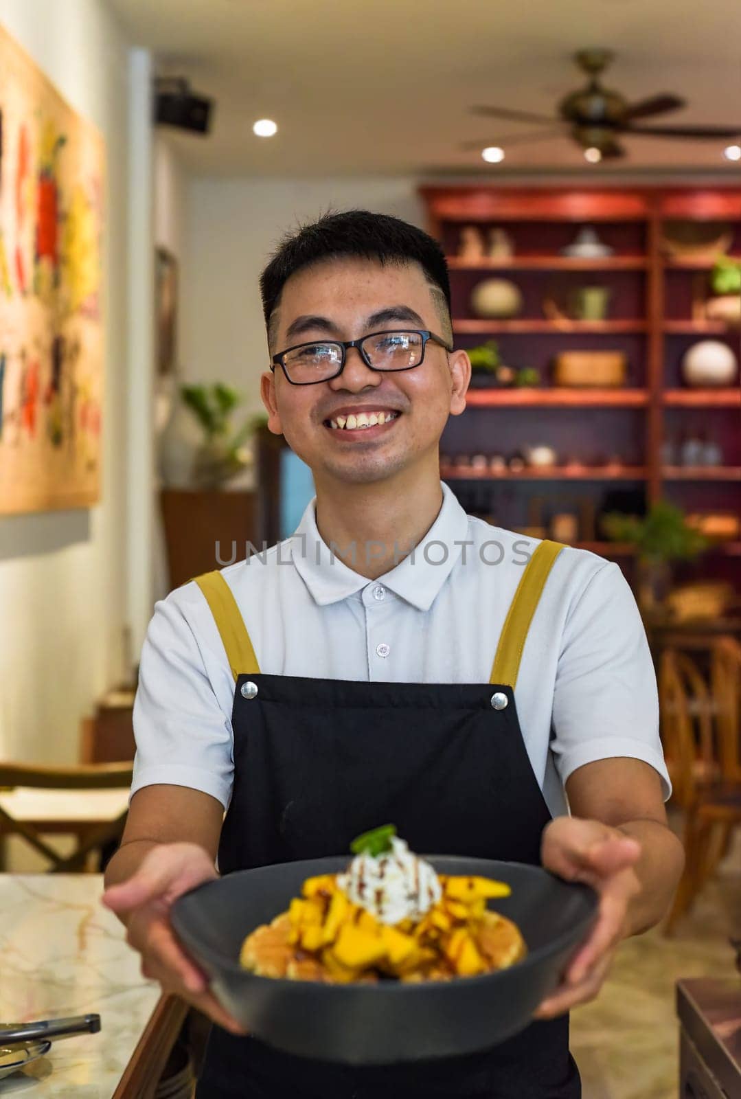 Vietnamese man chef in uniform holding fresh baked belgian waffles with cream and sliced mango fruit in cafe