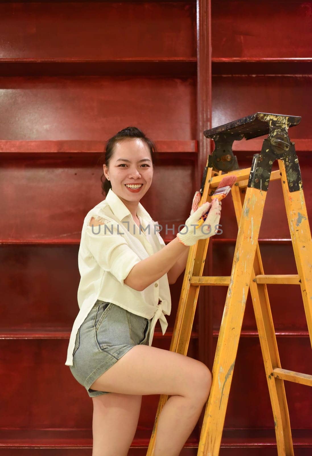 Vietnamese asian woman stands on a stepladder with a brush in her hand and paints the wall, makes repairs