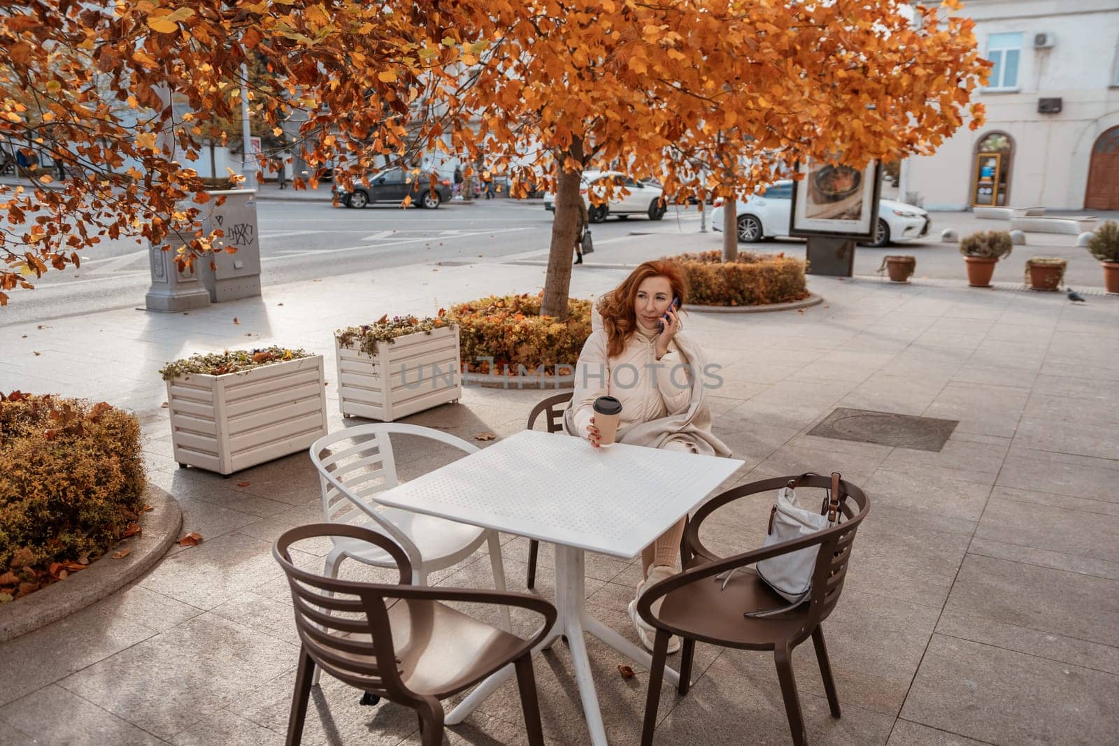 Woman sits at a white table outside a cafe under a vibrant autumn tree. She holds a cup; planters and chairs surround her.