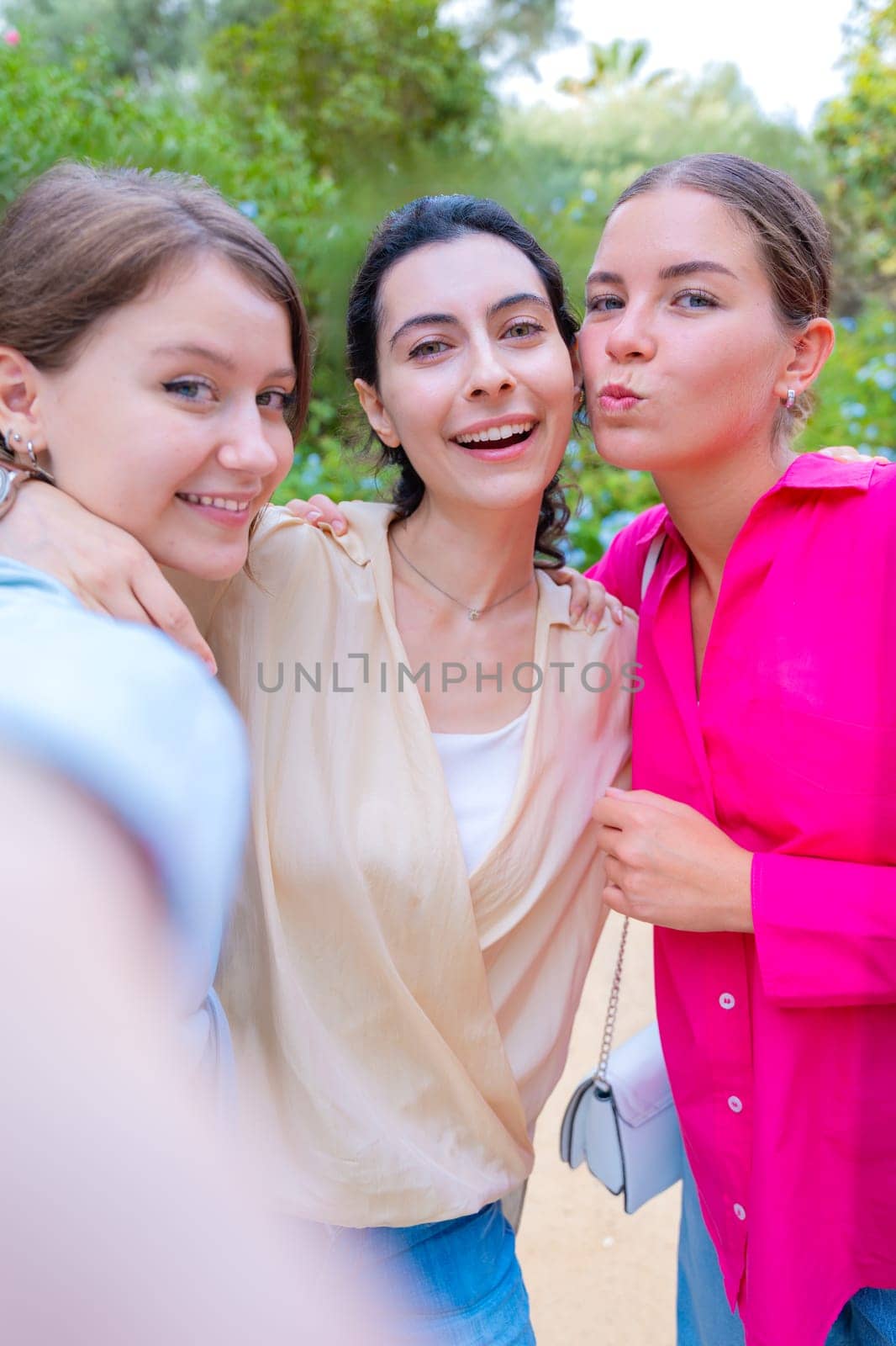 Three smiling women posing for a selfie in a lush green park. They embrace each other, expressing happiness and friendship. The vibrant colors and natural background create a joyful atmosphere.