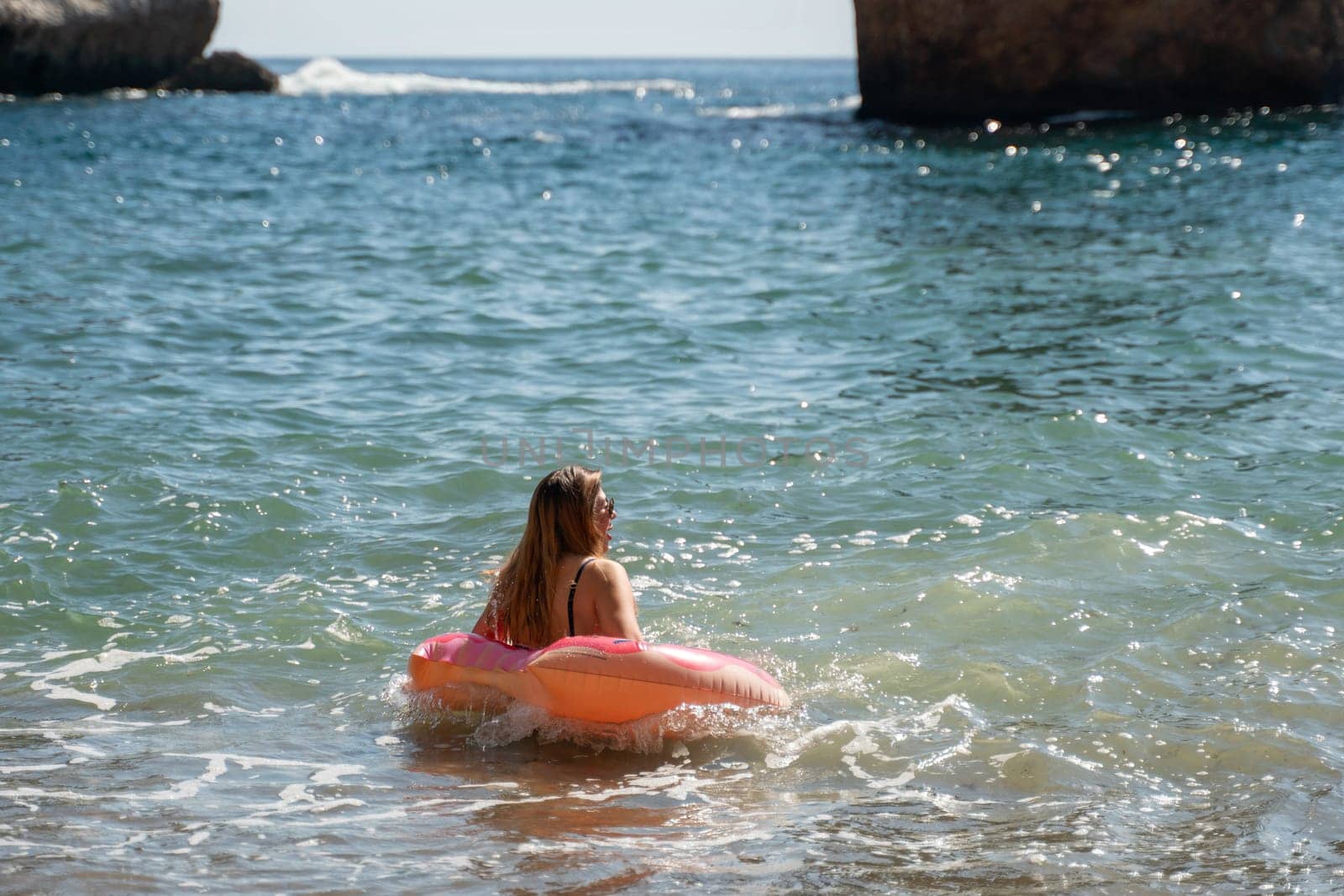 A woman floats on a pink inflatable ring in the ocean, facing away from the camera. Large rocks rise from the water in the background.