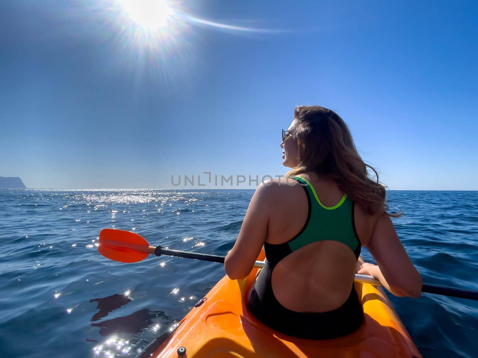 A woman in a green swimsuit paddles a yellow kayak on a calm ocean, with the sun shining brightly in the sky.