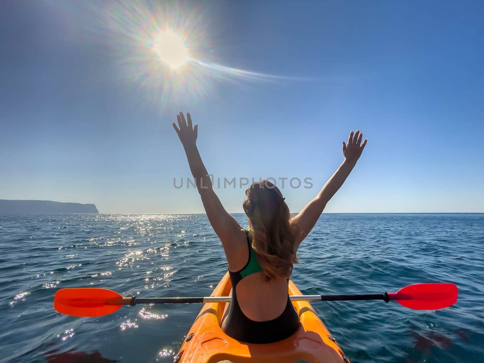 A woman in a swimsuit paddles a yellow kayak in the ocean, arms raised in the air, with the sun shining overhead.