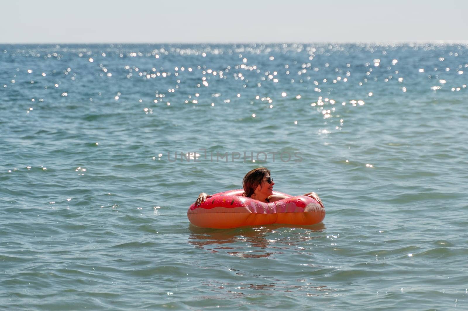 A person floats in a bright orange inflatable ring in a blue ocean with a bright sun reflecting off the water.