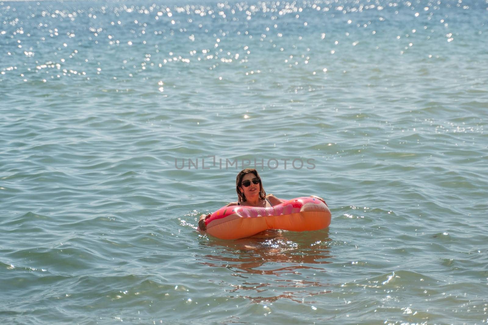 A woman floats in a bright orange inflatable ring in calm blue water, a sunny day at the beach.