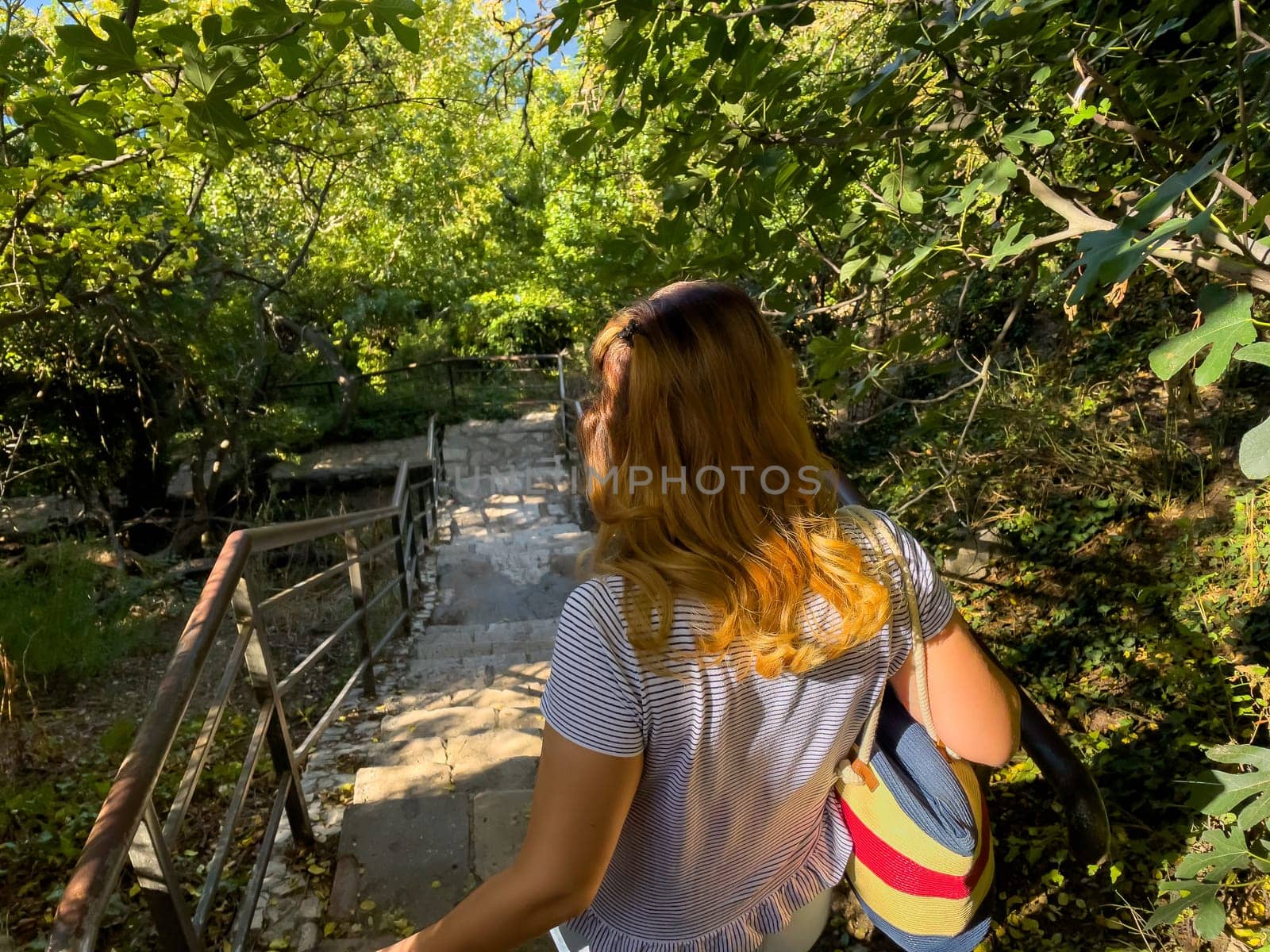 A woman with long blonde hair walks up a stone staircase with a handrail, surrounded by lush greenery. She is wearing a white t-shirt and carrying a blue and red bag.