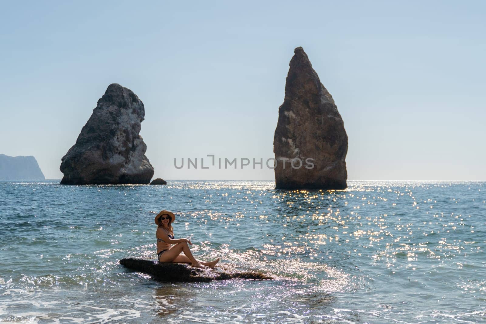 A woman sits on a rock in the water, facing two large rock formations, a calm sea stretches out behind.