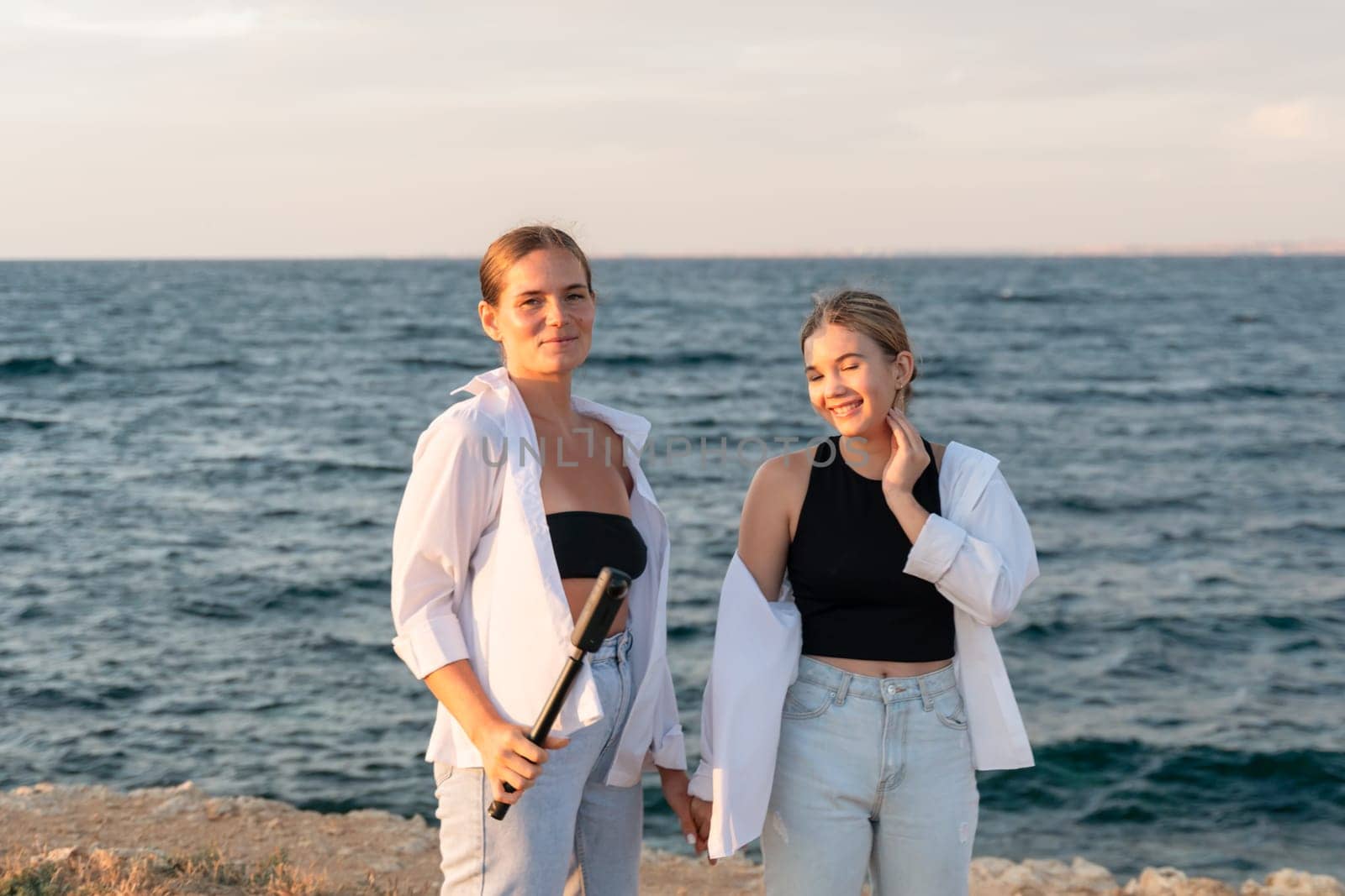 Two women stand on a rocky beach, looking at the camera. The woman on the left holds a white flute, the other woman has her hand on her cheek. The ocean is behind them.